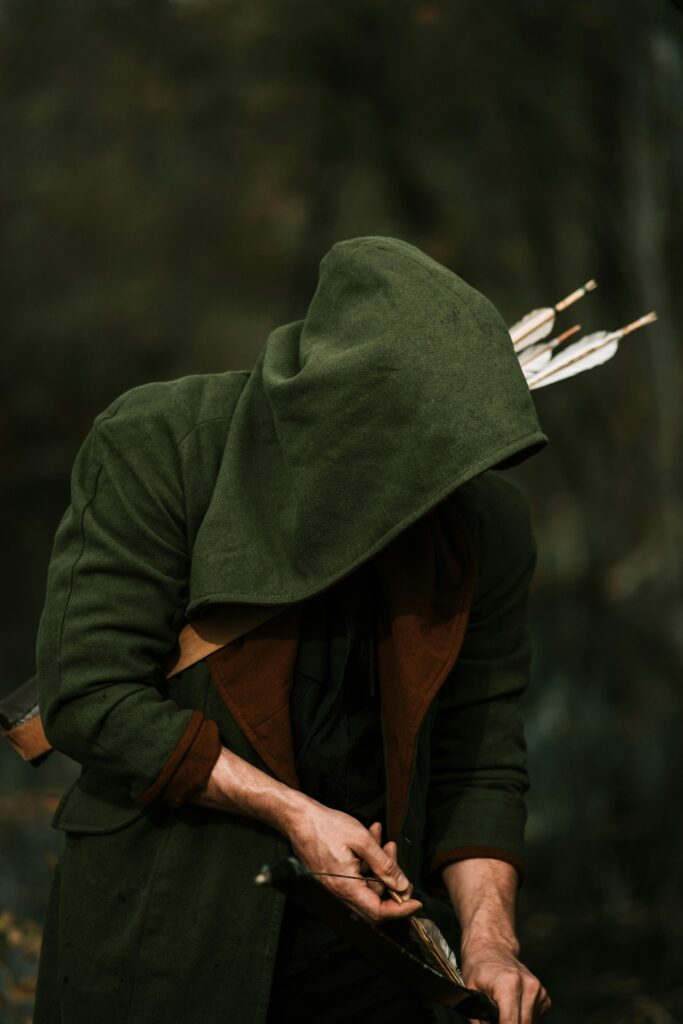 A hooded archer preparing arrows in a dense forest, embodying a hunting scene.